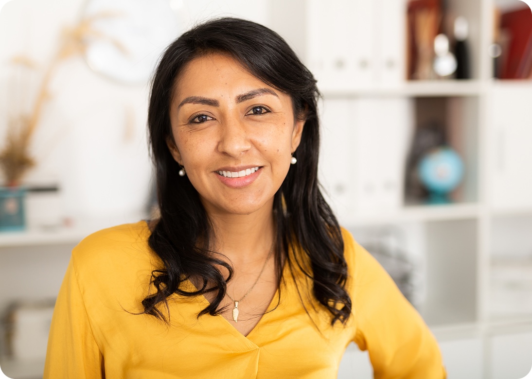 A woman with long dark hair, wearing a yellow blouse and a necklace, stands indoors smiling at the camera. The background shows white shelves with books, decor, and a globe.