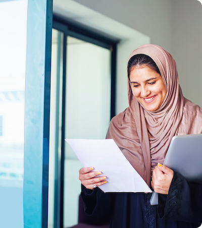 A woman wearing a brown hijab smiles while holding a document in one hand and a laptop in the other, standing indoors near a glass door.