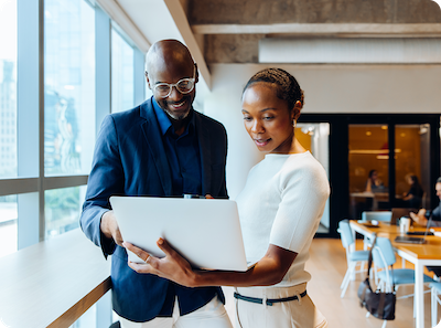 Two professionally dressed people stand by a window in a modern office, looking at a laptop together and smiling, with desks and chairs visible in the background.