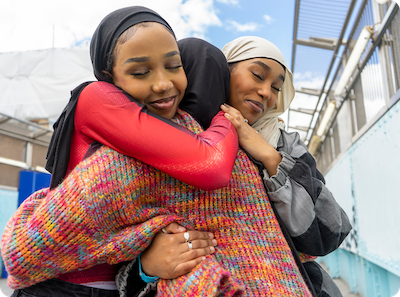 Three women wearing colorful hijabs and cozy clothing embrace each other in a warm group hug outdoors, smiling with eyes closed, with blue sky and buildings in the background.