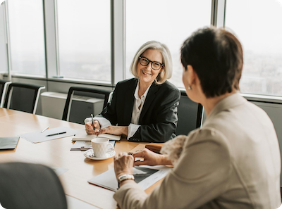 Two women sit at a conference table in an office, engaged in a discussion. One woman is smiling and holding a pen, while the other woman listens. Papers, notebooks, and a cup of coffee are on the table.