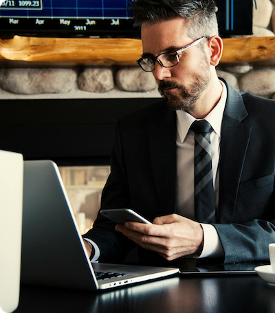 A man in a suit and glasses sits at a desk using a smartphone and a laptop, with a cup and financial chart visible in the background.