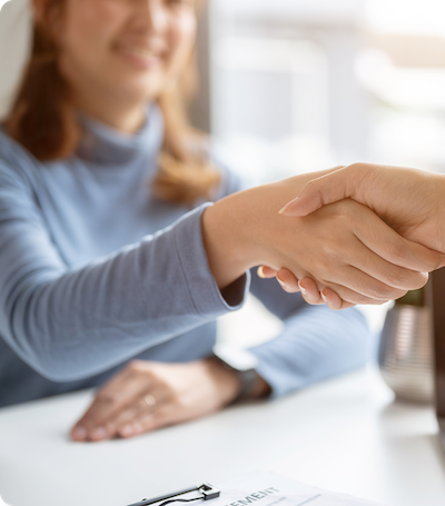 A person in a blue sweater smiles and shakes hands with another individual across a desk, suggesting a friendly meeting or agreement. A clipboard with paperwork is visible in the foreground.